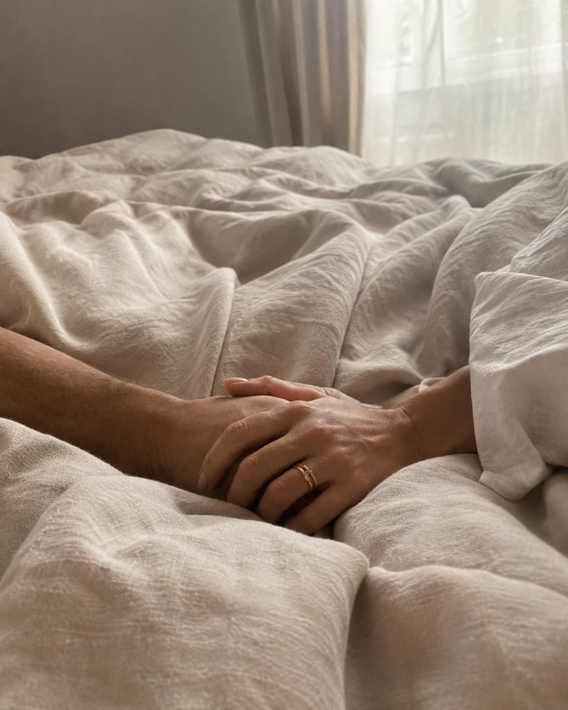 Two hands gently intertwined on rumpled cream linen bedding with soft window light, evoking intimacy and hormone wellness.